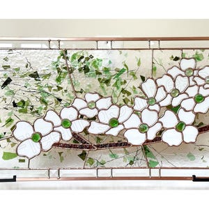 May include: A rectangular stained glass panel featuring white dogwood flowers with green glass centers on a brown branch. The background consists of clear textured glass with scattered green glass fragments, all encased in a copper metal frame.