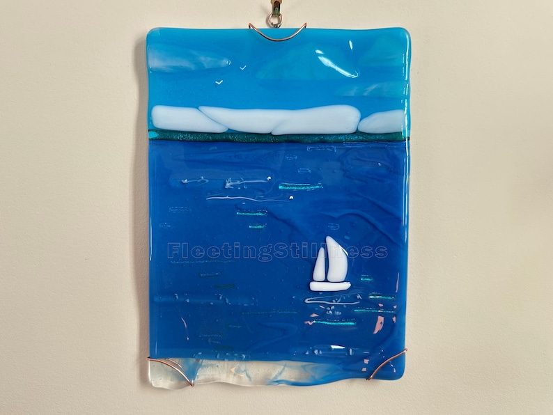 May include: A rectangular fused glass wall hanging featuring a blue sea and sky landscape. A small white sailboat floats on the water, with white clouds above the horizon. The text FleetingStillness is visible in the center of the glass piece.
