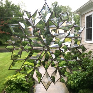 May include: A large, intricate, mirrored snowflake made of glass and metal. The snowflake is hanging in front of a house and reflects the surrounding greenery.