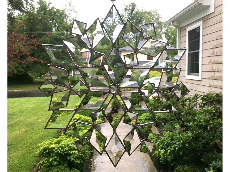 May include: A large, intricate, mirrored snowflake made of glass and metal. The snowflake is hanging in front of a house and reflects the surrounding greenery.