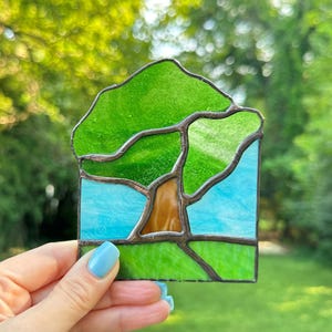 May include: A stained glass suncatcher featuring a tree with a brown trunk and a vibrant green canopy. The background consists of a light blue sky above a bright green grassy field. The entire scene is framed by dark metal solder lines.