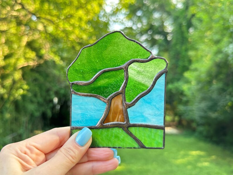 May include: A stained glass suncatcher featuring a tree with a brown trunk and a vibrant green canopy. The background consists of a light blue sky above a bright green grassy field. The entire scene is framed by dark metal solder lines.
