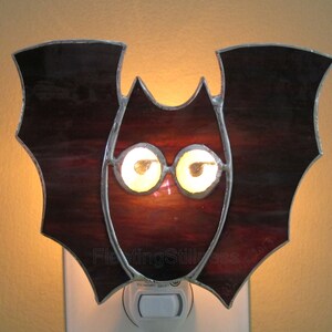 May include: A stained glass bat night light with a dark red body and two glowing yellow eyes. The night light is plugged into a standard electrical outlet.