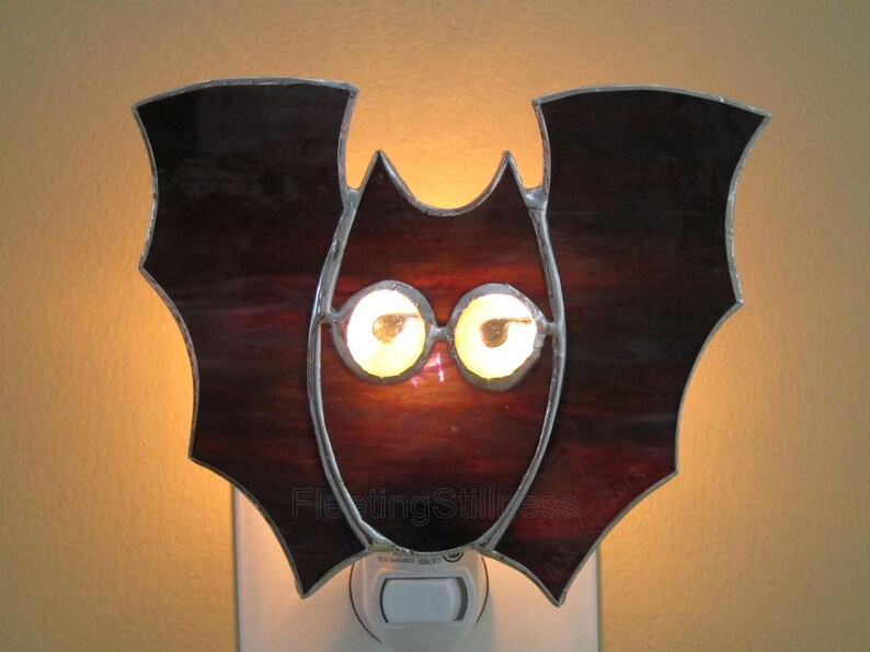 May include: A stained glass bat night light with a dark red body and two glowing yellow eyes. The night light is plugged into a standard electrical outlet.