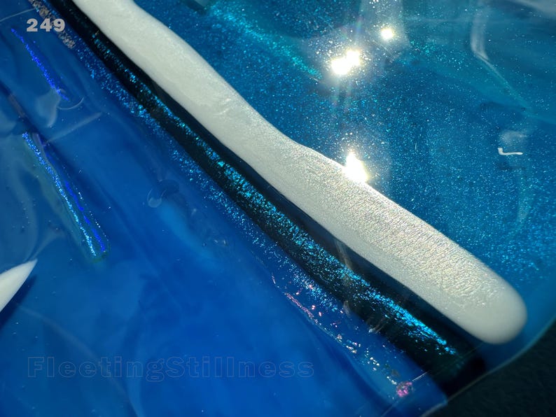 May include: A glossy blue resin surface featuring a prominent white curved ridge and a dark black line. The number 249 is printed in the top left corner, and the text FleetingStillness is visible in the bottom left area of the blue background.