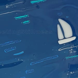 May include: A deep blue glass art piece featuring a white sailboat silhouette and scattered horizontal teal accents. The number 248 is printed in the top left corner, and the text FleetingStillness is visible in the center of the blue surface.