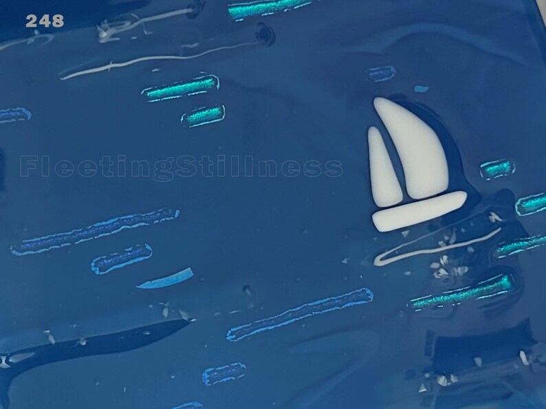 May include: A deep blue glass art piece featuring a white sailboat silhouette and scattered horizontal teal accents. The number 248 is printed in the top left corner, and the text FleetingStillness is visible in the center of the blue surface.