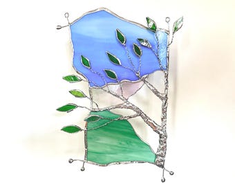 Op de afbeelding: Gebrandschilderd glaspaneel met een gestileerde boom met groene bladeren en takken. De achtergrond bevat blauw en groen glas, met een vleugje roze en wit. Het paneel is omlijst met zilverkleurig metaal.