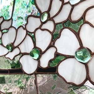 May include: A stained glass panel featuring white flower petals outlined in dark metal solder lines. Each flower center is accented with a smooth, translucent green glass gem. The background consists of various shades of green glass pieces.