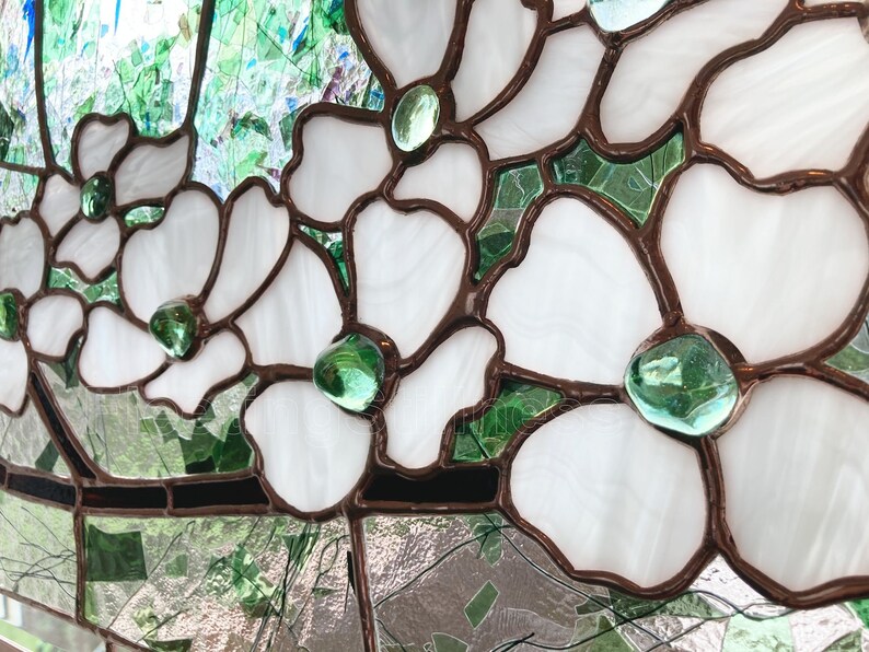 May include: A stained glass panel featuring white flower petals outlined in dark metal solder lines. Each flower center is accented with a smooth, translucent green glass gem. The background consists of various shades of green glass pieces.