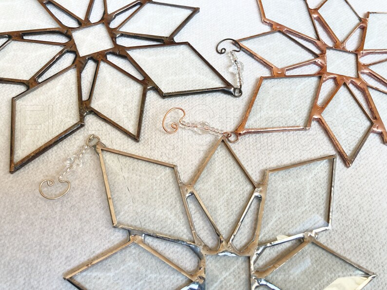 May include: Three stained glass snowflake ornaments with a diamond-shaped design. The snowflakes are made with clear glass and have a copper, silver, and gold finish.