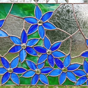 May include: A stained glass panel featuring multiple six-petaled blue flowers with yellow glass centers. The flowers are connected by copper-colored metal solder lines and set against a background of textured clear and solid green glass pieces.