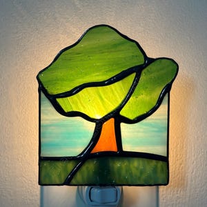 May include: A square stained glass night light featuring a tree design. The tree has a bright orange trunk and vibrant green glass leaves. The background consists of light blue and white glass panels, while the base is a solid dark green glass.