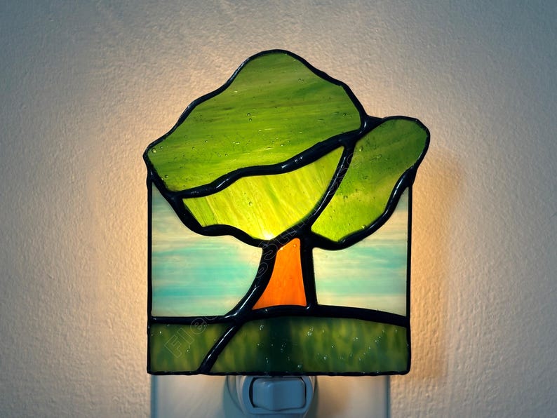 May include: A square stained glass night light featuring a tree design. The tree has a bright orange trunk and vibrant green glass leaves. The background consists of light blue and white glass panels, while the base is a solid dark green glass.