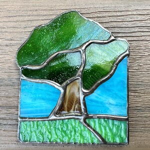 May include: A stained glass suncatcher featuring a tree with a brown trunk and green canopy set against a bright blue sky. The base of the piece shows a textured light green grassy area. The entire design is held together by dark metal solder lines.
