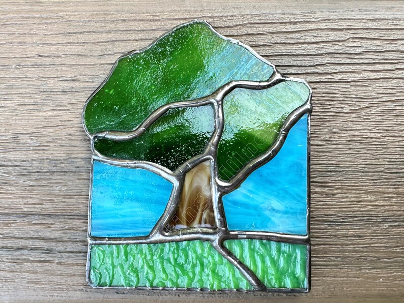 May include: A stained glass suncatcher featuring a tree with a brown trunk and green canopy set against a bright blue sky. The base of the piece shows a textured light green grassy area. The entire design is held together by dark metal solder lines.