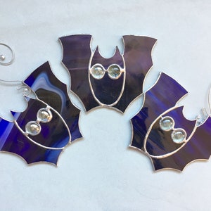 May include: Three purple stained glass bats with silver wire accents and clear glass eyes. The bats are hanging from silver wire loops.