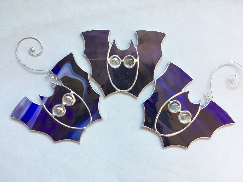 May include: Three purple stained glass bats with silver wire accents and clear glass eyes. The bats are hanging from silver wire loops.