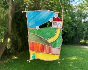 Op de afbeelding: Een glas-in-lood zonnevanger met een landschapstafereel met een blauwe lucht, een wit huis met een rood dak, groene heuvels, een geel pad en een gele voorgrond. Een kleine blauwe uilfiguur is onderaan bevestigd, samen met witte bloemaccenten.