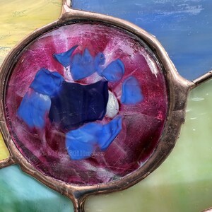 K&ouml;nnte beinhalten: Ein kreisf&ouml;rmiges Buntglasfenster mit einem tief magentafarbenen Zentrum, das mit verschiedenen blauen Glassplittern besetzt ist. Das St&uuml;ck ist mit Kupferfolien-L&ouml;tlinien eingefasst und von gelben, blauen und gr&uuml;nen Glassegmenten umgeben.