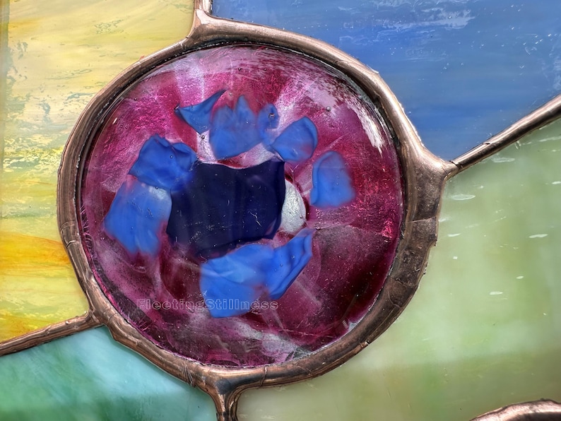 K&ouml;nnte beinhalten: Ein kreisf&ouml;rmiges Buntglasfenster mit einem tief magentafarbenen Zentrum, das mit verschiedenen blauen Glassplittern besetzt ist. Das St&uuml;ck ist mit Kupferfolien-L&ouml;tlinien eingefasst und von gelben, blauen und gr&uuml;nen Glassegmenten umgeben.