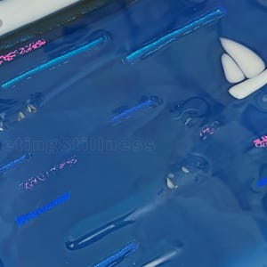 May include: A glossy blue resin surface featuring embedded white sailboat shapes, shimmering pink and blue glitter accents, and the text 249 and FleetingStillness. The composition displays a smooth, reflective finish with various linear and abstract elements.