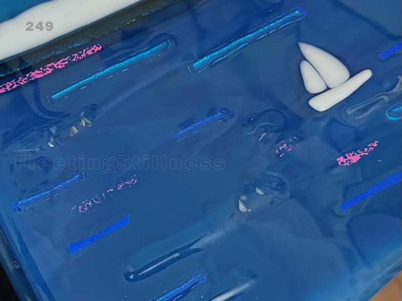 May include: A glossy blue resin surface featuring embedded white sailboat shapes, shimmering pink and blue glitter accents, and the text 249 and FleetingStillness. The composition displays a smooth, reflective finish with various linear and abstract elements.