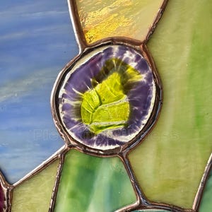 K&ouml;nnte beinhalten: Ein Buntglasfenster mit einem zentralen kreisf&ouml;rmigen Glaselement, das einen violetten Rand und ein leuchtend lindgr&uuml;nes Zentrum aufweist. Die umliegenden Glassegmente sind in Blau-, Gelb- und Gr&uuml;nt&ouml;nen gehalten und durch d&uuml;nne kupferfarbene Metalll&ouml;tstellen verbunden.