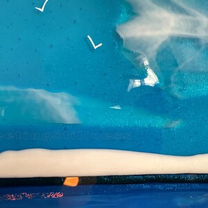 May include: A fused glass art piece featuring a vibrant blue sky with white bird silhouettes and wispy white clouds. The lower section displays a solid white horizontal band above a dark blue base. The text 249 and FleetingStillness are visible.