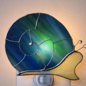 May include: A stained glass nightlight shaped like a snail with a blue and green shell and a yellow body. The nightlight is plugged into an outlet.