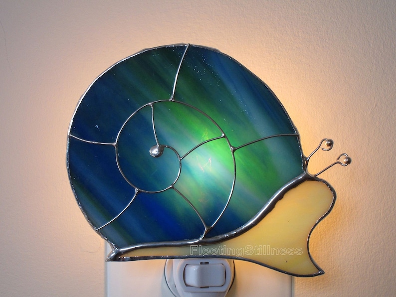 May include: A stained glass nightlight shaped like a snail with a blue and green shell and a yellow body. The nightlight is plugged into an outlet.