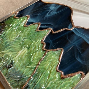 May include: A stained glass artwork depicting a mountain range with green, blue, and white glass pieces. The glass is textured and has a metallic finish.