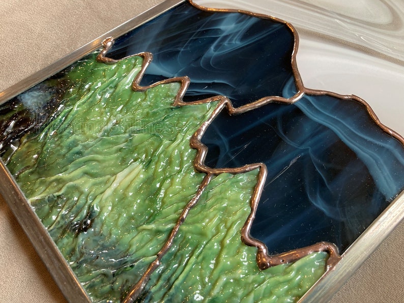 May include: A stained glass artwork depicting a mountain range with green, blue, and white glass pieces. The glass is textured and has a metallic finish.