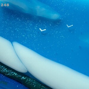 May include: A rectangular fused glass piece featuring a vibrant blue background with embedded glitter, two smooth white curved shapes, and the number 248 printed in the top left corner. The surface contains small air bubbles and a dark green strip.