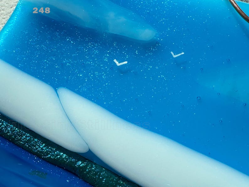 May include: A rectangular fused glass piece featuring a vibrant blue background with embedded glitter, two smooth white curved shapes, and the number 248 printed in the top left corner. The surface contains small air bubbles and a dark green strip.