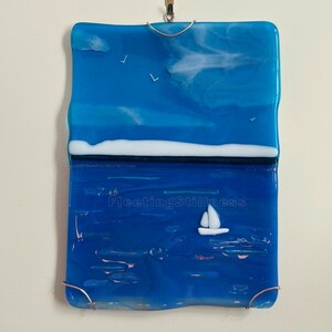 May include: A rectangular fused glass wall hanging featuring a seascape with a bright blue sky, white clouds, and three small birds. A white horizontal line separates the sky from a deep blue sea with a white sailboat and the text FleetingStillness.