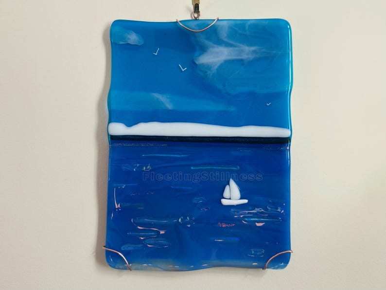 May include: A rectangular fused glass wall hanging featuring a seascape with a bright blue sky, white clouds, and three small birds. A white horizontal line separates the sky from a deep blue sea with a white sailboat and the text FleetingStillness.