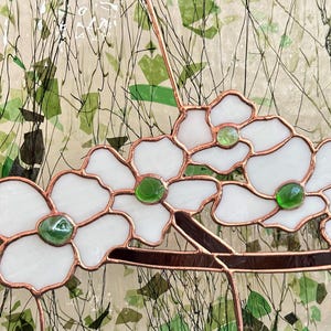 May include: A stained glass panel featuring white flower petals with copper foil edges and small, round, translucent green glass centers. The flowers are attached to a dark brown glass branch, set against a textured, light-colored background.