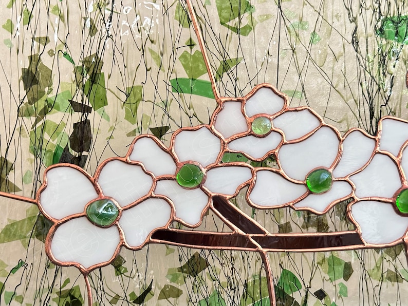 May include: A stained glass panel featuring white flower petals with copper foil edges and small, round, translucent green glass centers. The flowers are attached to a dark brown glass branch, set against a textured, light-colored background.