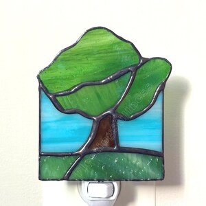 May include: A stained glass night light featuring a tree with a brown trunk and green canopy set against a light blue sky and a green grassy foreground. The piece is constructed with dark metal solder lines and includes the text floatingstillness.