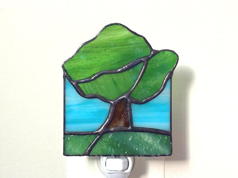 May include: A stained glass night light featuring a tree with a brown trunk and green canopy set against a light blue sky and a green grassy foreground. The piece is constructed with dark metal solder lines and includes the text floatingstillness.