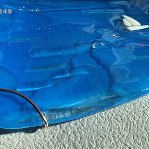 May include: A rectangular blue glass tray featuring a white sailboat graphic, iridescent accents, and the text 249 and FleetingStillness. A curved copper wire element is attached to the bottom left corner of the smooth, translucent glass surface.