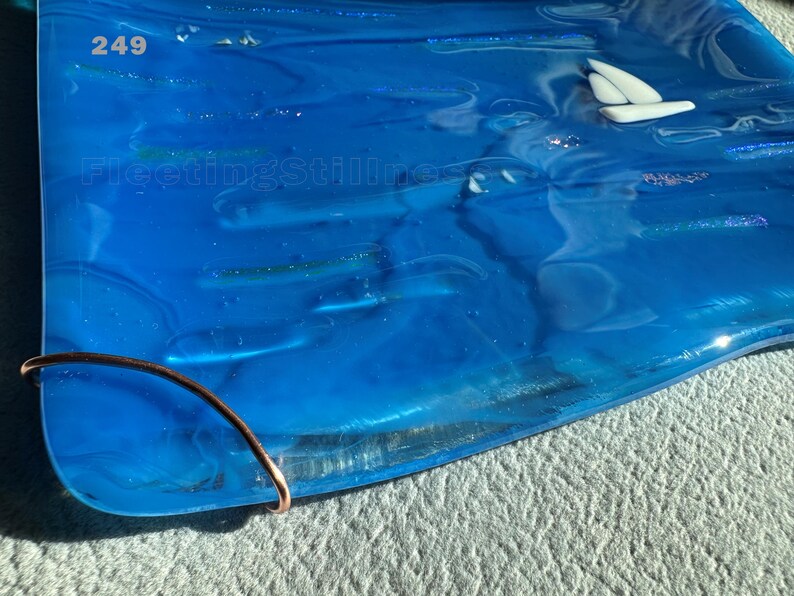 May include: A rectangular blue glass tray featuring a white sailboat graphic, iridescent accents, and the text 249 and FleetingStillness. A curved copper wire element is attached to the bottom left corner of the smooth, translucent glass surface.