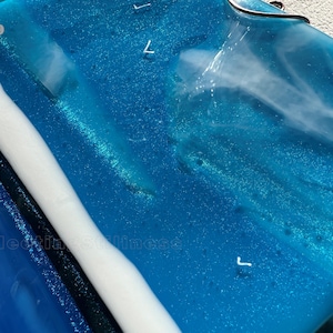 May include: A rectangular blue glass piece with a glossy finish, featuring swirling white opaque accents and embedded glitter. The number 249 is printed in the top left corner, and the text Fleeting Stillness is visible in the lower left area.