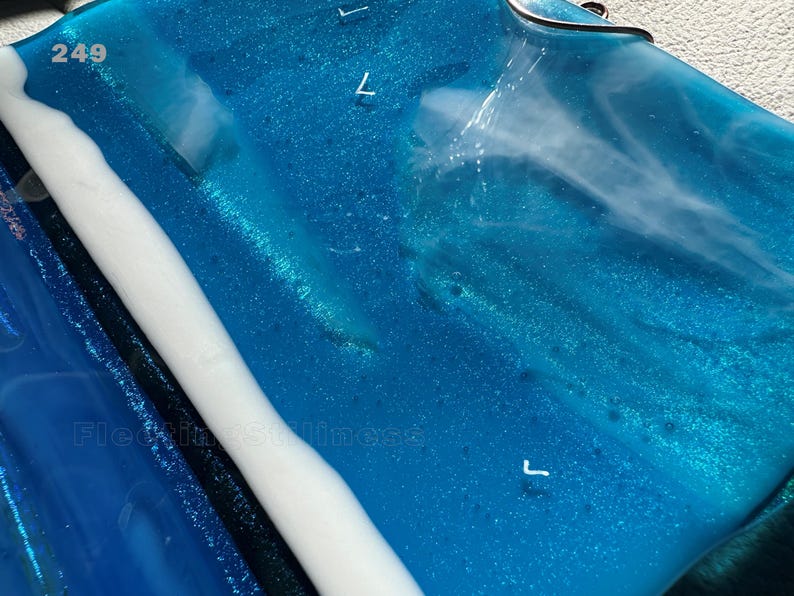 May include: A rectangular blue glass piece with a glossy finish, featuring swirling white opaque accents and embedded glitter. The number 249 is printed in the top left corner, and the text Fleeting Stillness is visible in the lower left area.