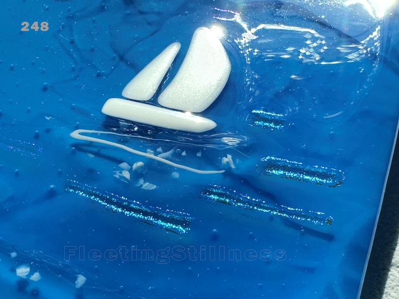 May include: A glossy blue resin art piece featuring a white sailboat silhouette with a shimmering finish. The surface displays the number 248 in the top left corner and the text FleetingStillness at the bottom, accented by blue glitter streaks.