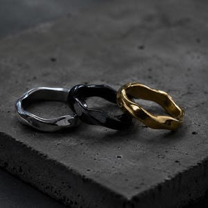 Peut inclure: Trois bagues en argent, noir et or, chacune avec un design texturé et organique. Les bagues sont présentées sur une surface grise foncée et texturée, mettant en valeur leurs formes uniques et irrégulières. La bague dorée a une finition brillante.