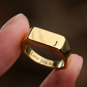 May include: A gold-colored signet ring with a rectangular face and rounded edges. The letter "J" is engraved on the face. The words "Love you dad" are engraved on the inside of the ring.