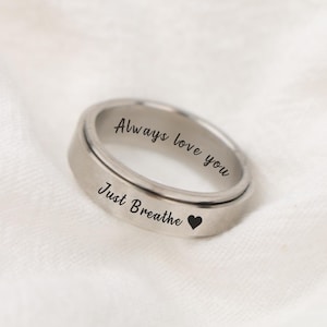 May include: A silver-colored ring with the words "Always love you" and "Just Breathe" engraved on the outer surface. A small black heart symbol is next to the words "Just Breathe". The ring is set against a white, textured background.