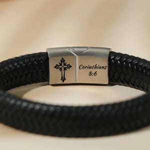 May include: Black braided leather bracelet with a silver clasp. The clasp features a cross design and the text "Corinthians 8:6". The bracelet is a religious accessory.